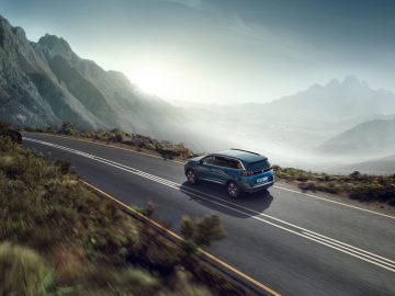 Een blauwe Peugeot 5008 SUV rijdt op een bochtige bergweg met dramatische rotspieken en zonlicht op de achtergrond.