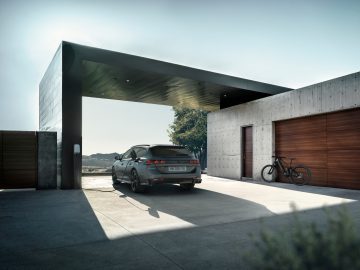 Een elektrische Peugeot 508 Peugeot Sport Engineered laadt op in de strakke carport van een modern huis, met betonnen muren en een houten garagedeur, te midden van een serene natuurlijke omgeving.