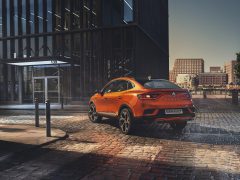Oranje Renault Arkana geparkeerd op een geplaveide straat in een stedelijke omgeving met moderne gebouwen en een helderblauwe lucht.