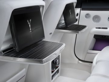 Interieur van een luxe Rolls-Royce Ghost-auto met twee gevouwen dienbladtafels met schermen, met het logo in reliëf op één, afgezet tegen witleren stoelen.