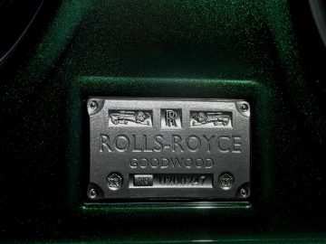 Close-up van een metalen Rolls-Royce Ghost-naamplaatje op een groene, sprankelende achtergrond, gemarkeerd met "Goodwood" en een serienummer.