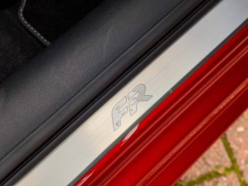 Close-up van de deurdrempel van een Seat Leon Sportstourer met een metalen logo, afgezet tegen een rood autoframe en een zwart lederen interieur.
