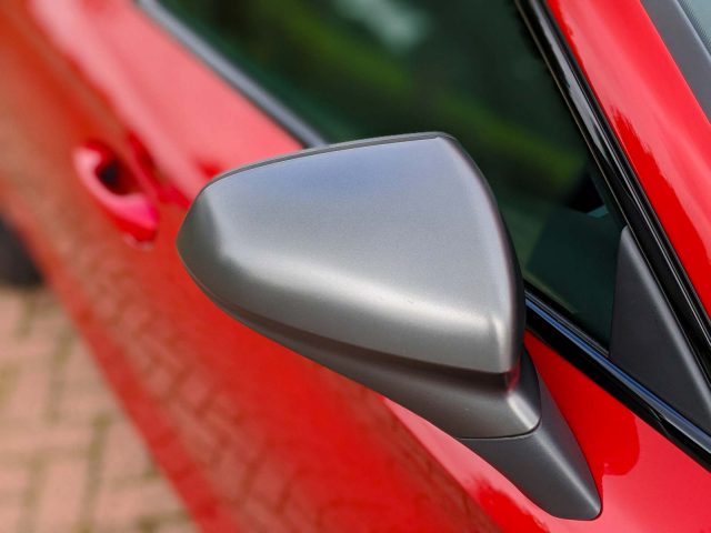 Close-up van een matgrijze zijspiegel van een rode Seat Leon Sportstourer, waarbij de details en textuur van de spiegelbehuizing centraal staan.