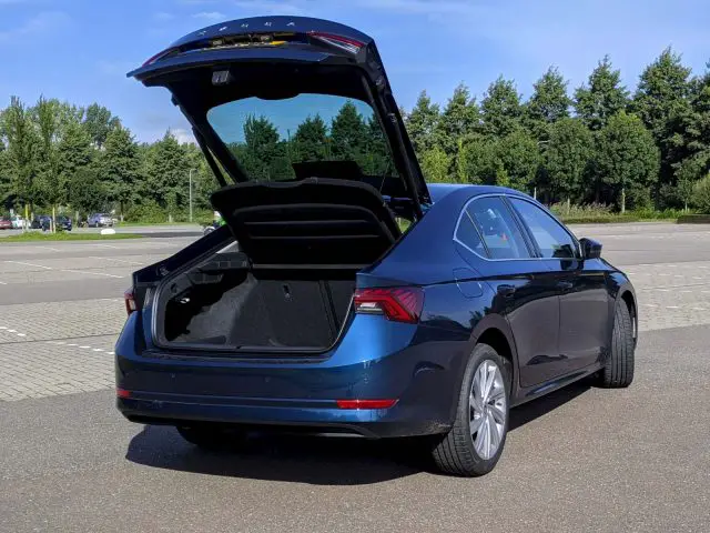 Een blauwe Skoda Octavia sedan met open kofferbak, geparkeerd op een zonnige parkeerplaats met bomen op de achtergrond.