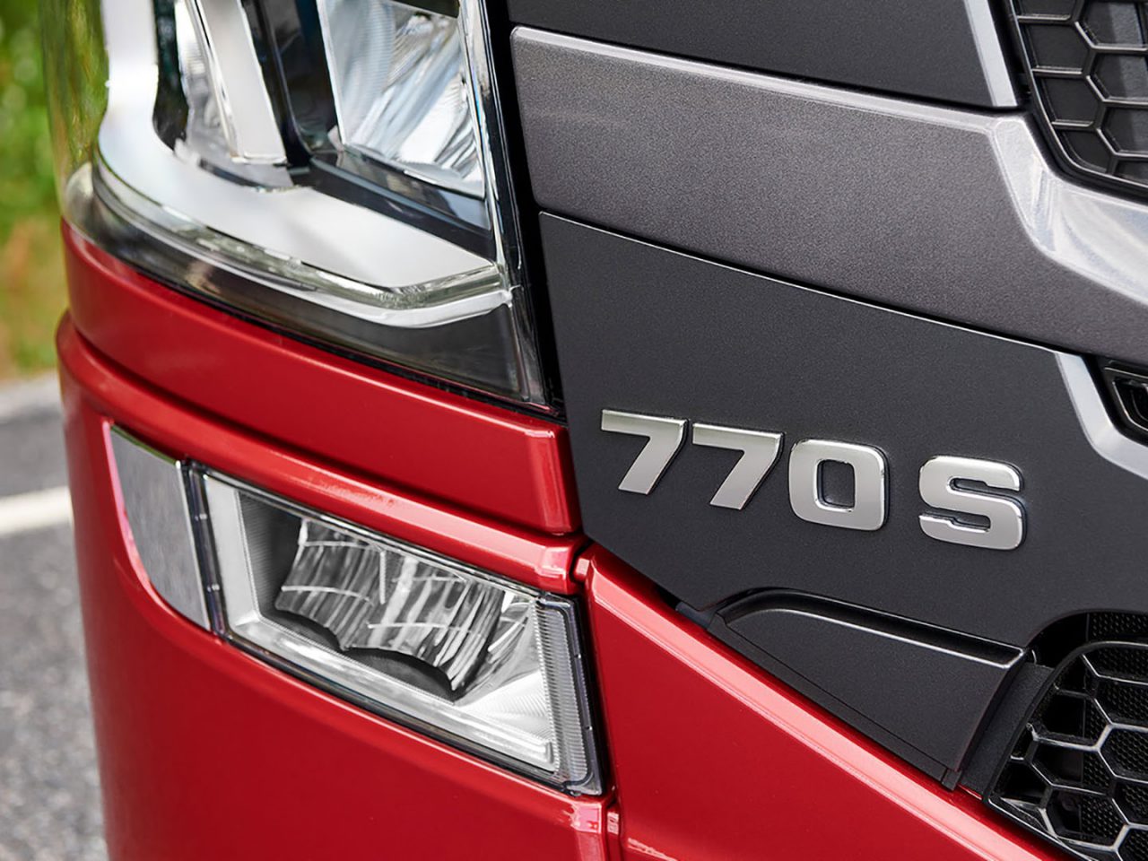 Close-up van de voorkant van een rode Scania V8-truck met het modelnummer "770s" op de grille, met gedetailleerde accenten op de koplamp en het grillepatroon.