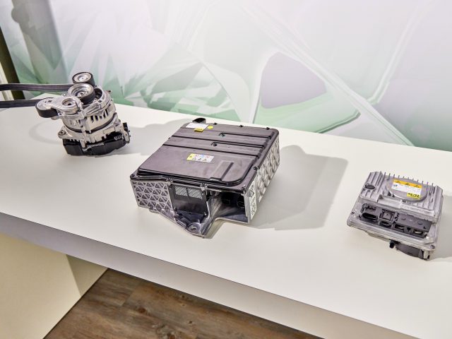 Drie auto-onderdelen van een Skoda Octavia iV weergegeven op een wit oppervlak: een mechanische riemaandrijving aan de linkerkant, een grote elektronische regeleenheid in het midden en een kleinere module aan de linkerkant.