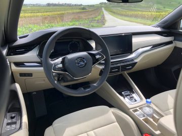 Binnenaanzicht van een Skoda Octavia iV met stuur, dashboard en crèmekleurige lederen stoelen, uitkijkend op een landelijke weg en wijngaard.