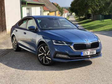 Een blauwe Skoda Octavia iV sedan geparkeerd op een dorpsweg met huizen en groene heuvels op de achtergrond op een zonnige dag.