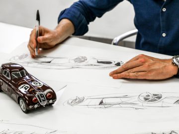 Een persoon die aan een bureau ontwerpen schetst van een Touring AERO 3-auto, met een modelauto en verschillende tekeningen eromheen verspreid.