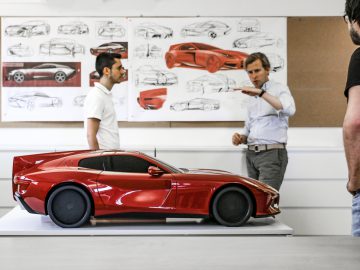 Twee mannen bespreken een Touring AERO 3-model in een ontwerpstudio met autoschetsen aan de muur.