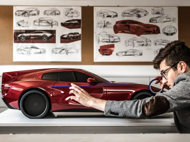 Een mannelijke ontwerper onderzoekt een schaalmodel van de rode sportwagen "Touring AERO 3" met ontwerpschetsen aan de muur achter hem.