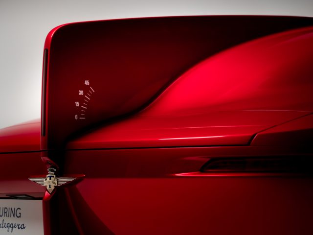 Close-up van de achterkant van een rode toersportwagen, met de nadruk op de AERO 3-vleugel en het embleem met zichtbare snelheidsmarkeringen.