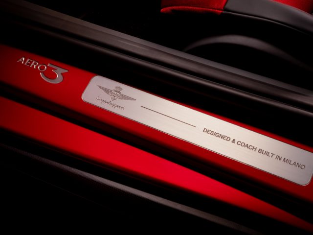 Close-up van een metalen naamplaatje op een rood oppervlak met de inscriptie "Touring AERO 3", een logo en de tekst "ontworpen door een in Milaan gebouwde touringcar.