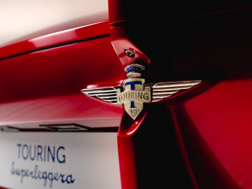 Close-up van het Touring AERO 3-embleem op een rode auto met een onscherpe achtergrond met de woorden 'touring superleggera'.