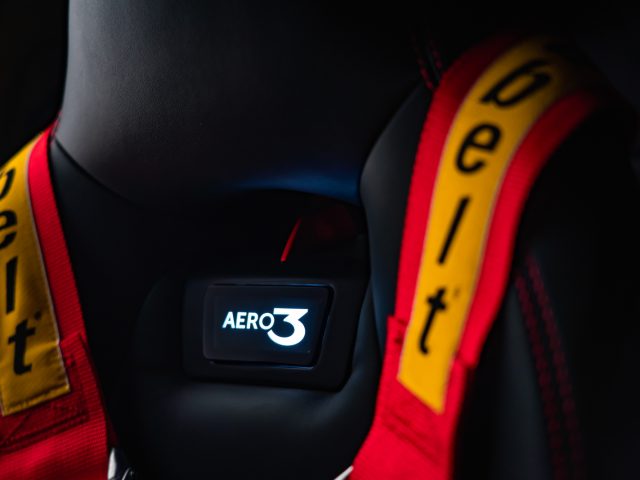 Close-up van een autostoeltje met een zwart en rood ontwerp, met opvallende gele veiligheidsgordels gemarkeerd met "pelt" en een stoelgesp met de tekst "Touring AERO 3".