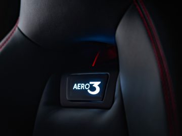 Close-up van de hoofdsteun van een autostoel met een verlicht 'Touring AERO 3'-logo, met zwarte bekleding en rode stiksels.