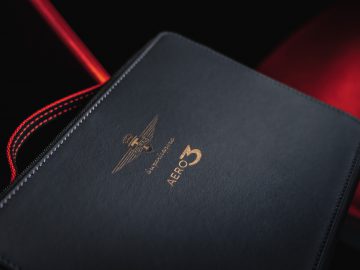 Een zwart notitieboekje met "Touring AERO 3" en een gouden vliegtuiglogo in reliëf op de omslag, rustend op een rode en zwarte achtergrond.