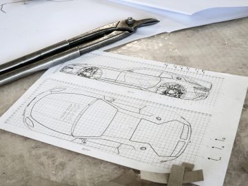 Technische tekeningen van het Touring AERO 3-autoontwerp op ruitjespapier met een metalen schaalliniaal en een tang op een werkplaatstafel.