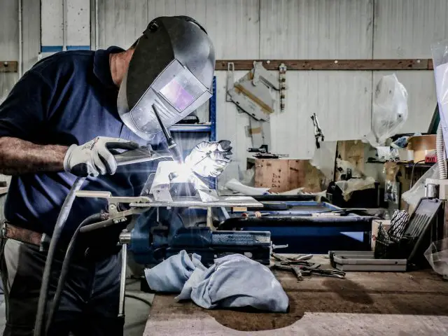 Een arbeider in beschermende uitrusting gebruikt een Touring AERO 3-lasapparaat op een metalen stuk in een industriële werkplaats.