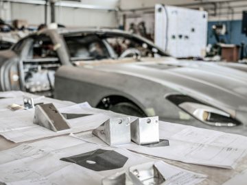 Auto-onderdelen en technische tekeningen op een werkbank in een autoreparatiewerkplaats, terwijl op de achtergrond een Touring AERO 3 wordt onderhouden.
