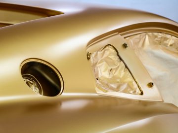 Close-up van een auto in een werkplaats, met details van een gedeeltelijk gemaskeerde Touring AERO 3 met een gouden afwerking, waarbij de nadruk ligt op het koplampgebied.