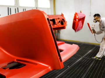 Werknemer in beschermende kleding spuit auto-onderdelen in een werkplaats, met focus op een rode Touring AERO 3-autobumper op de voorgrond.