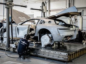 Een technicus werkt aan een gedemonteerde Touring AERO 3-sportwagen in een drukke garage, waar in de ruimte diverse gereedschappen en apparatuur zichtbaar zijn.