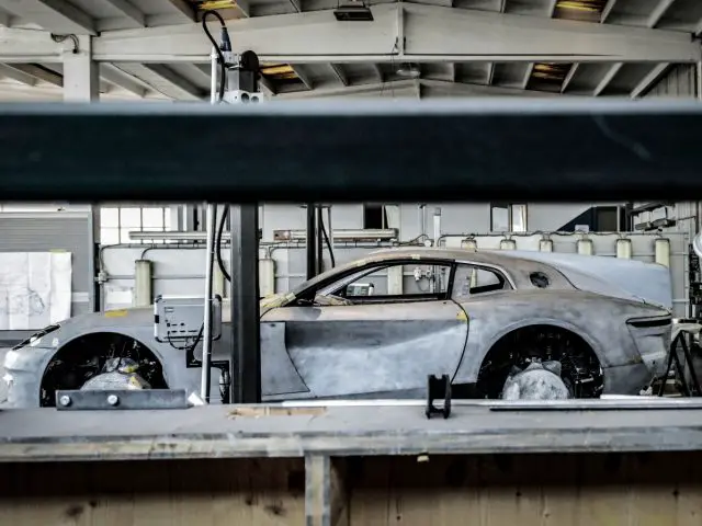 Een gedeeltelijk geassembleerde Touring AERO 3-sportwagen in een werkplaats, bekeken door een open raam, met rondom zichtbaar gereedschap en uitrusting.