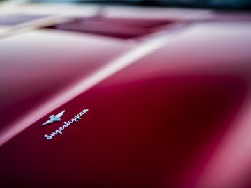 Close-up van de motorkap van een rode auto met een chroom "Touring AERO 3" embleem en vage lamellen op de achtergrond.
