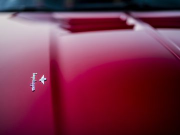 Close-up van een levendig rode motorkap van een Touring AERO 3-sportwagen met een glanzend metallic logo, met de nadruk op strakke designlijnen en glanzende afwerking.