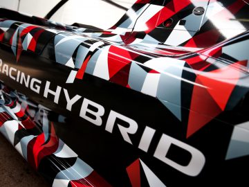 Close-up van het zijpaneel van een Toyota GR Super Sport hybride auto met een geometrisch rood, wit en zwart wrap-ontwerp en het woord "racinghybrid" prominent weergegeven.