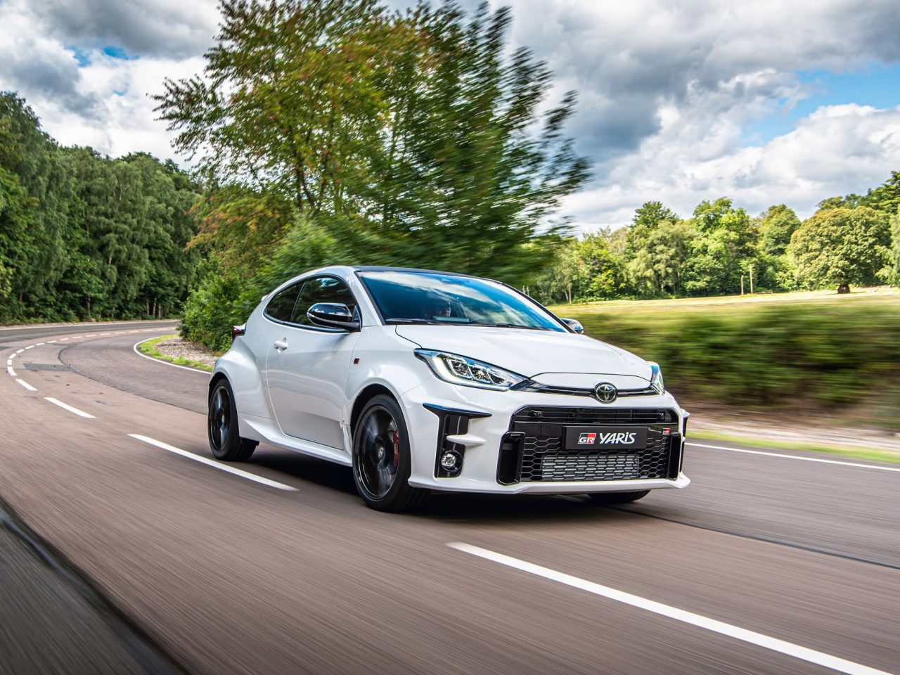 Een witte Toyota GR Yaris die met hoge snelheid over een gebogen bosweg rijdt onder een bewolkte hemel.