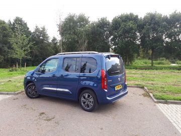 Een blauwe Toyota ProAce City Verso geparkeerd op een verhard terrein met gras en bomen op de achtergrond op een bewolkte dag.