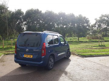 Blauwe Toyota ProAce City Verso bestelwagen geparkeerd op een verhard terrein met bomen en groen gras op de achtergrond op een zonnige dag.