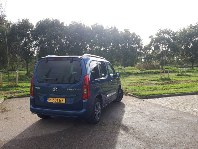 Blauwe Toyota ProAce City Verso bestelwagen geparkeerd op een grindterrein met groene velden en bomen op de achtergrond op een zonnige dag.