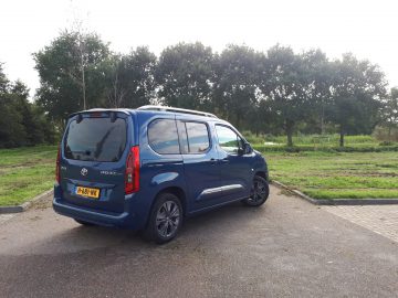 Blauwe Toyota ProAce City Verso bestelwagen geparkeerd op een verhard terrein met gras en bomen op de achtergrond op een bewolkte dag.