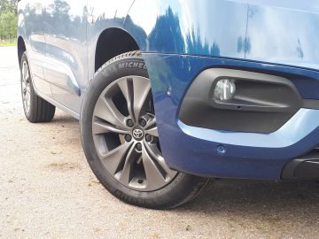 Close-up van de rechter voorband en bumper van een Toyota ProAce City Verso, met een schoon lichtmetalen velg en een Michelin-band geparkeerd op asfalt.