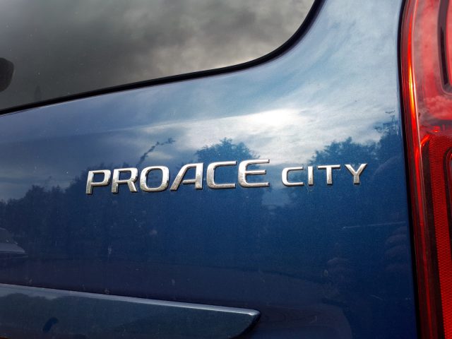 Close-up van het "Toyota ProAce City"-embleem op de achterkant van een voertuig, met een weerspiegeling van wolken en bomen op het blauwe oppervlak.