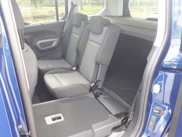 Binnenaanzicht van een Toyota ProAce City Verso met grijze stoffen achterbank en een open deur, met zichtbare veiligheidsgordels en vloervakken.