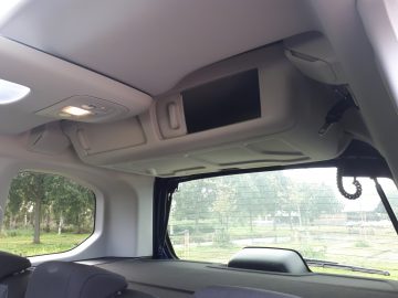 Binnenaanzicht van een Toyota ProAce City Verso met een open dakcompartiment met een scherm, omgeven door grijze bekleding, en een glimp van bomen door het zijraam.