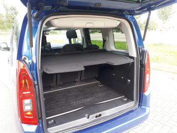 Open kofferbak van een blauwe Toyota ProAce City Verso met een lege laadruimte met neergeklapte stoelen, geparkeerd in een zonnige buitenruimte.