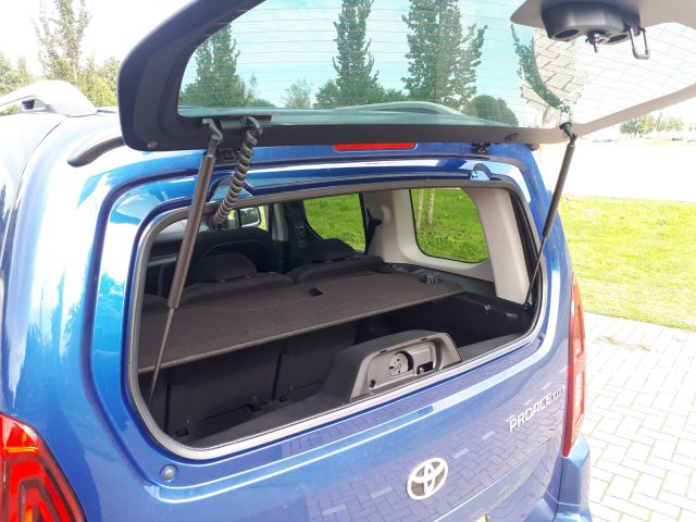 Achteraanzicht van een blauwe Toyota ProAce City Verso met een open kofferbak, met een neergeklapte achterbank om de laadruimte te vergroten, buiten geparkeerd.