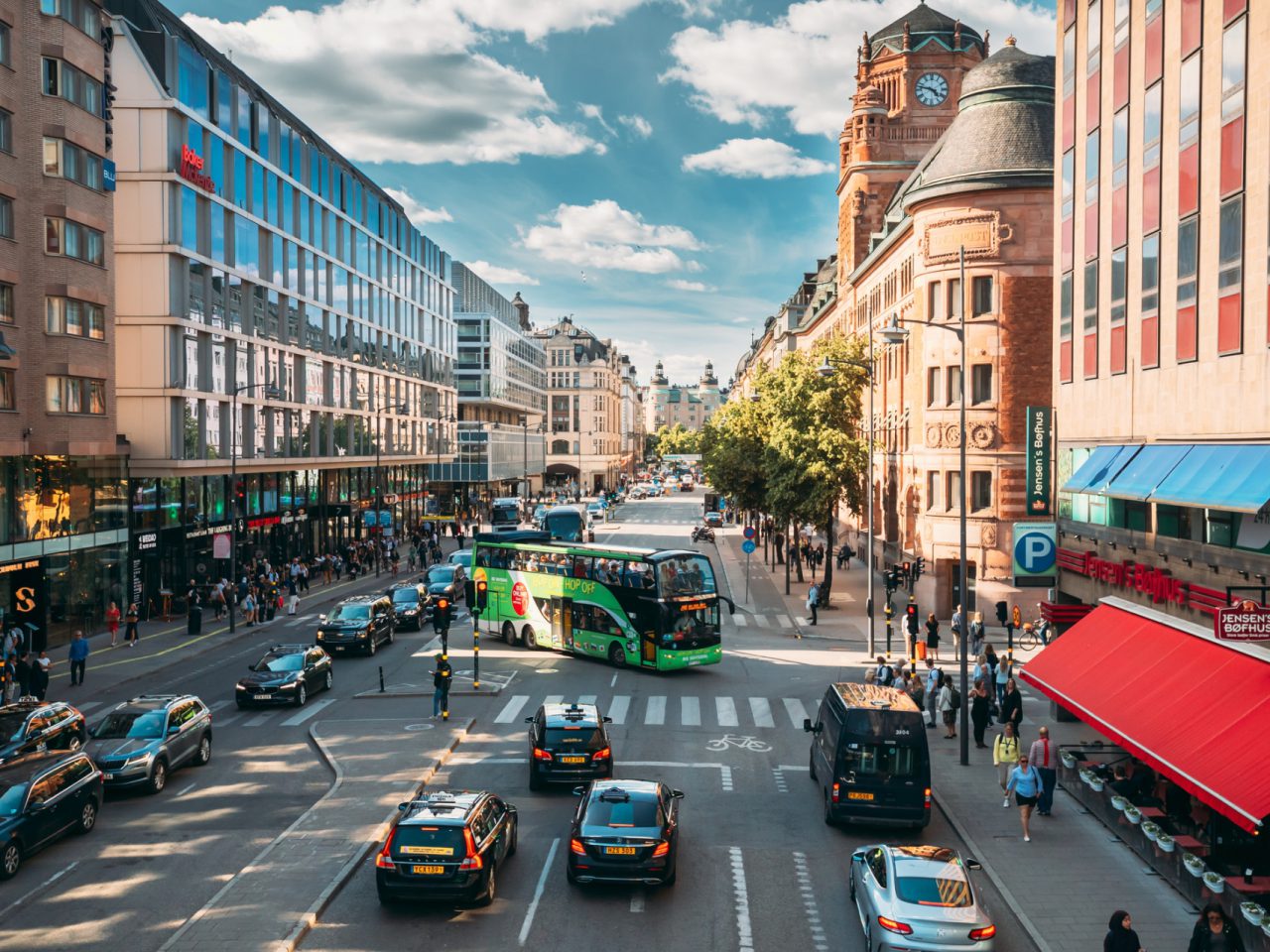 Een drukke stadsstraat met auto's, een bus en voetgangers die individuele mobiliteit beoefenen, geflankeerd door moderne en historische gebouwen op een zonnige dag.