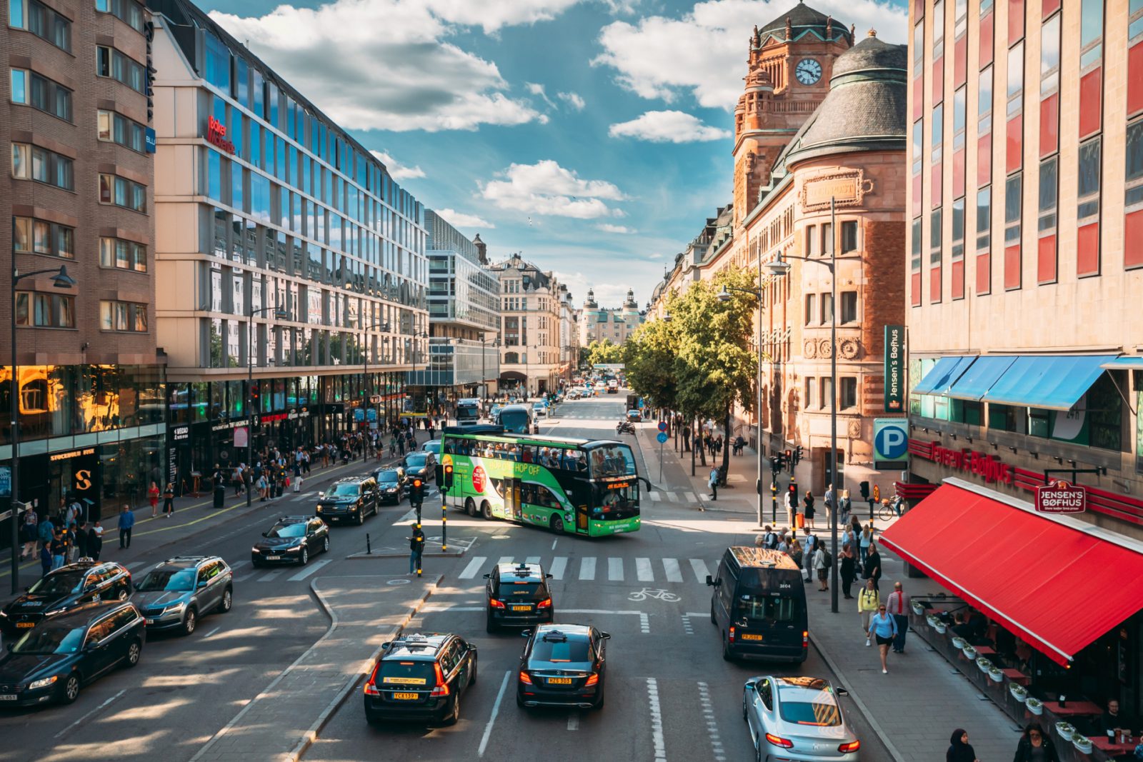 Een drukke stadsstraat met auto's, een bus en voetgangers die individuele mobiliteit beoefenen, geflankeerd door moderne en historische gebouwen op een zonnige dag.