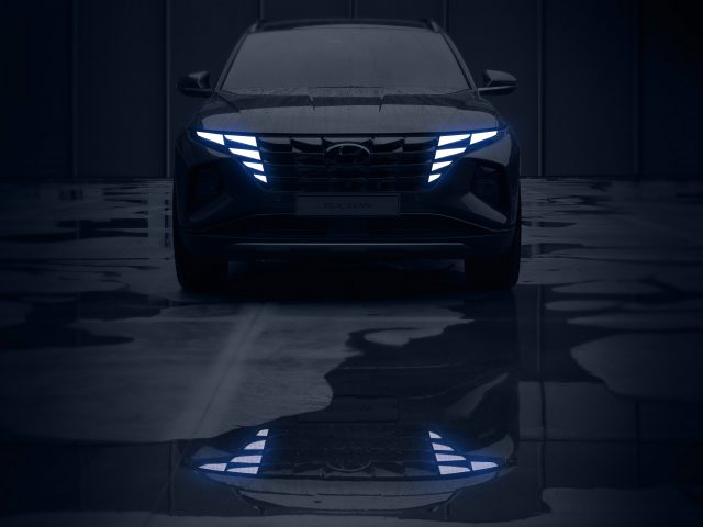Een geparkeerde Hyundai Tucson, met koplampen en grille verlicht in een blauwgekleurde omgeving, reflecterend op een nat oppervlak.