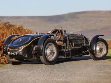 Een vintage zwarte Bugatti-raceauto geparkeerd op een landweg met heuvels op de achtergrond en gebladerte aan de zijkant.