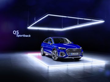 Een blauwe Audi Q5 Sportback tentoongesteld onder geometrische witte neonlichten in een donkere showroomomgeving.