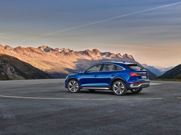 Een blauwe Audi Q5 Sportback geparkeerd op een brede weg met op de achtergrond zonovergoten bergketens onder een heldere hemel.
