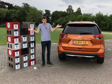 Man die tussen een hoge stapel dozen en een Nissan X-Trail staat en met een verwarde uitdrukking op een parkeerplaats gebaart.