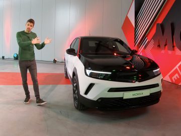 Man in groene trui presenteert een witte Opel Mokka-auto in een showroom met een rode en witte grafische achtergrond.